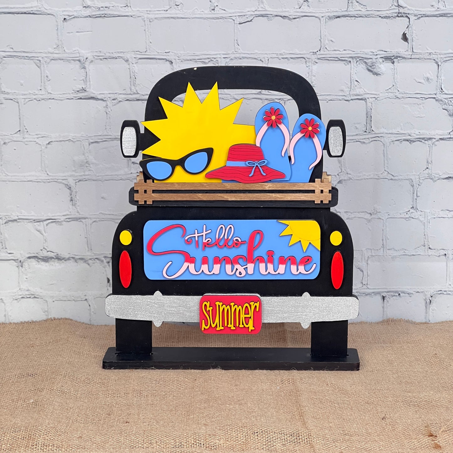 The Hello Sunshine Interchangeable Insert from Janet's Craft Corner is a DIY decor kit with 3 pieces shaped like a truck bed. It features "Hello Sunshine," "Summer," and sun, flip-flops, and sunhat designs. Ideal for summer picnics against a white brick wall backdrop.