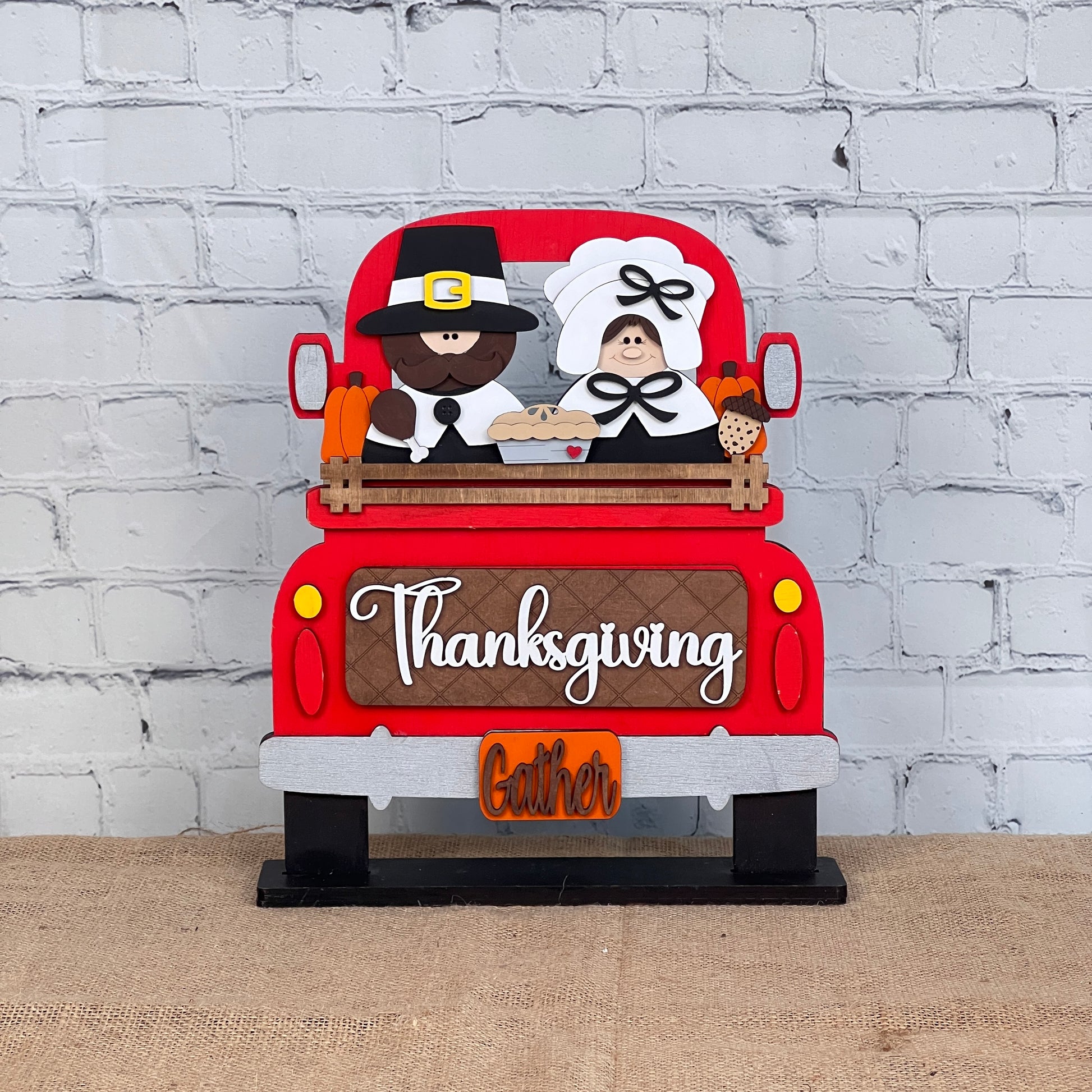 Janet's Craft Corner offers a Pilgrim Interchangeable Insert as part of their DIY home decor kit, featuring a red truck with a pilgrim couple, pie, and corn. "Thanksgiving" and "Gather" appear on the tailgate, set against a white brick wall—perfect for charming Thanksgiving decor.