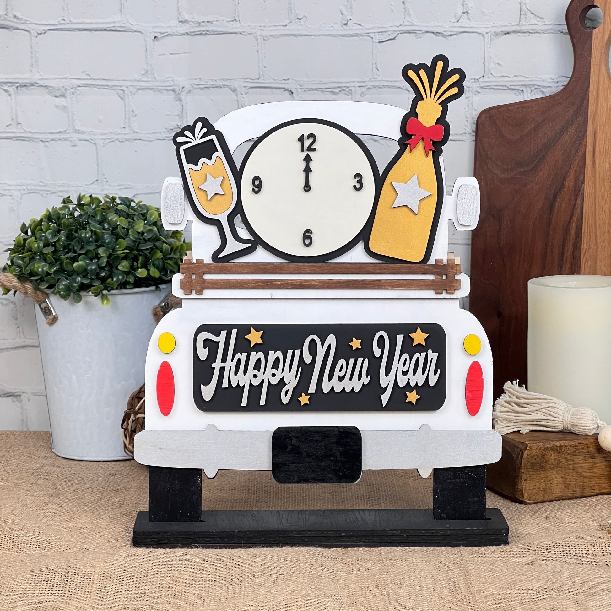 Unveil Janet's Craft Corner's DIY kit: an Antique Truck Base features "Happy New Year" on the back, adorned with a clock, champagne glass, and bottle. Perfect against a white brick wall with a potted plant and candle for versatile elegance.