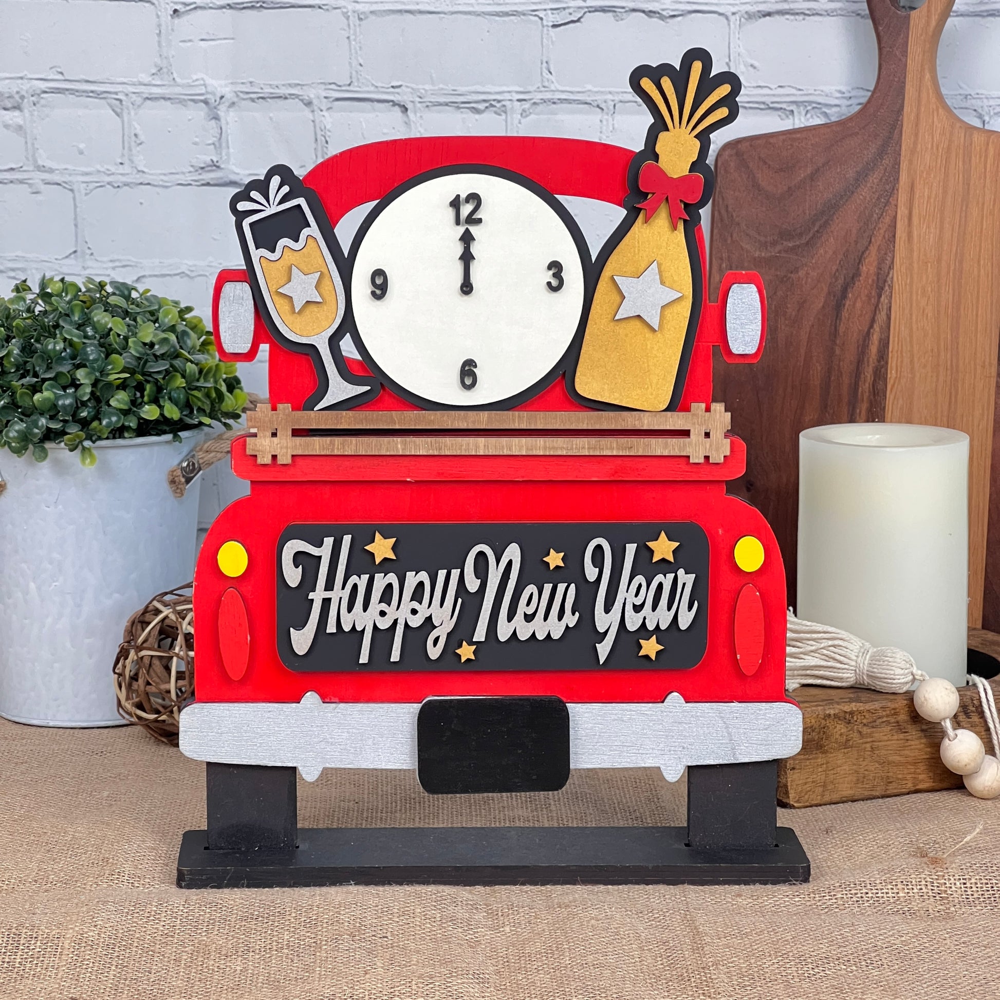 Janet's Craft Corner presents the New Year Interchangeable Insert, a DIY home decor kit with 3 pieces: a decorative red truck adorned with "Happy New Year," clock, champagne glass, and party popper. Perfect against a brick wall with plants and candles for festive charm.