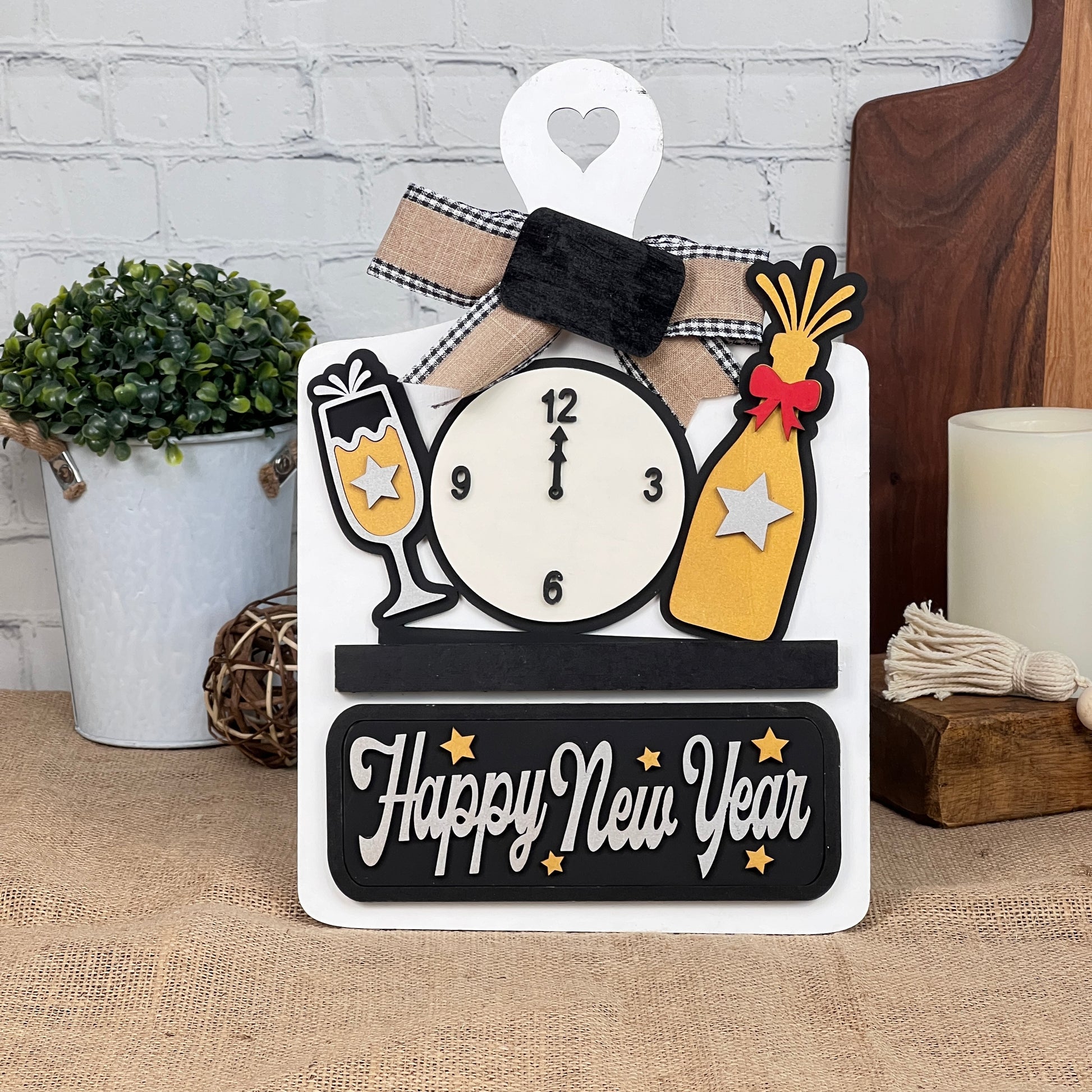 Janet's Craft Corner offers the New Year Interchangeable Insert DIY Kit, featuring a clock at midnight, champagne elements in black and gold, "Happy New Year" script, and background decor with a plant, candle, and wooden board. Perfect for seasonal home decoration.