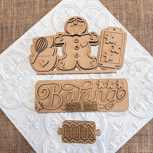 Wooden cookie-themed cutouts are set on a textured surface. The top cutout showcases a gingerbread figure along with baking tools, while the middle bears the phrase "Baking Spirits Bright." This DIY Craft Kit from Janet's Craft Corner is ideal for creating personalized home décor and features the Baking Spirits Bright Interchangeable Insert labeled "Cookies.