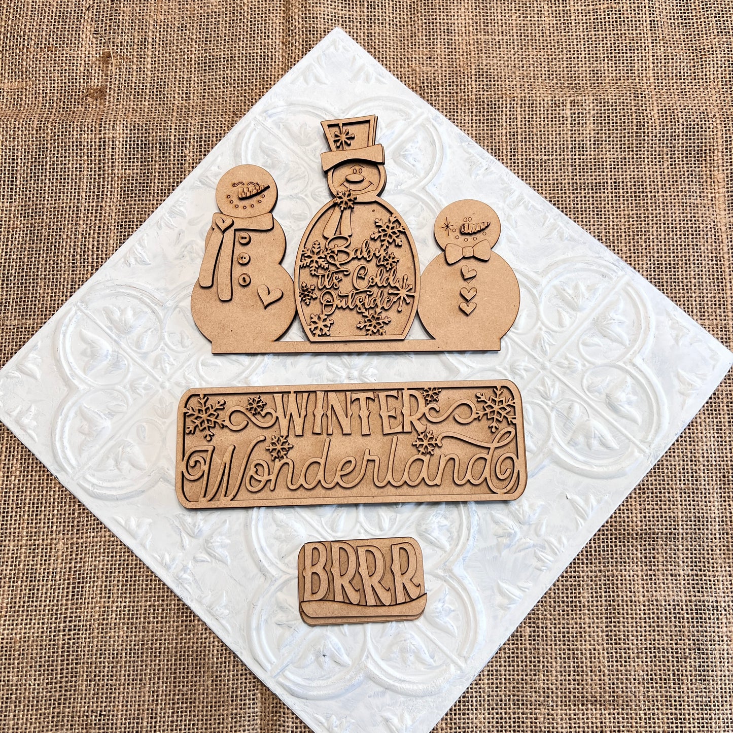 Wooden cutouts on a textured, white square background showcase two snowmen—one adorned with a top hat and scarf, the other smaller. The text "Winter Wonderland" and "BRRR" is displayed in decorative lettering. Ideal for personalized home décor, the burlap surface beneath adds a cozy touch. This is the Winter Wonderland Interchangeable Insert by Janet's Craft Corner.