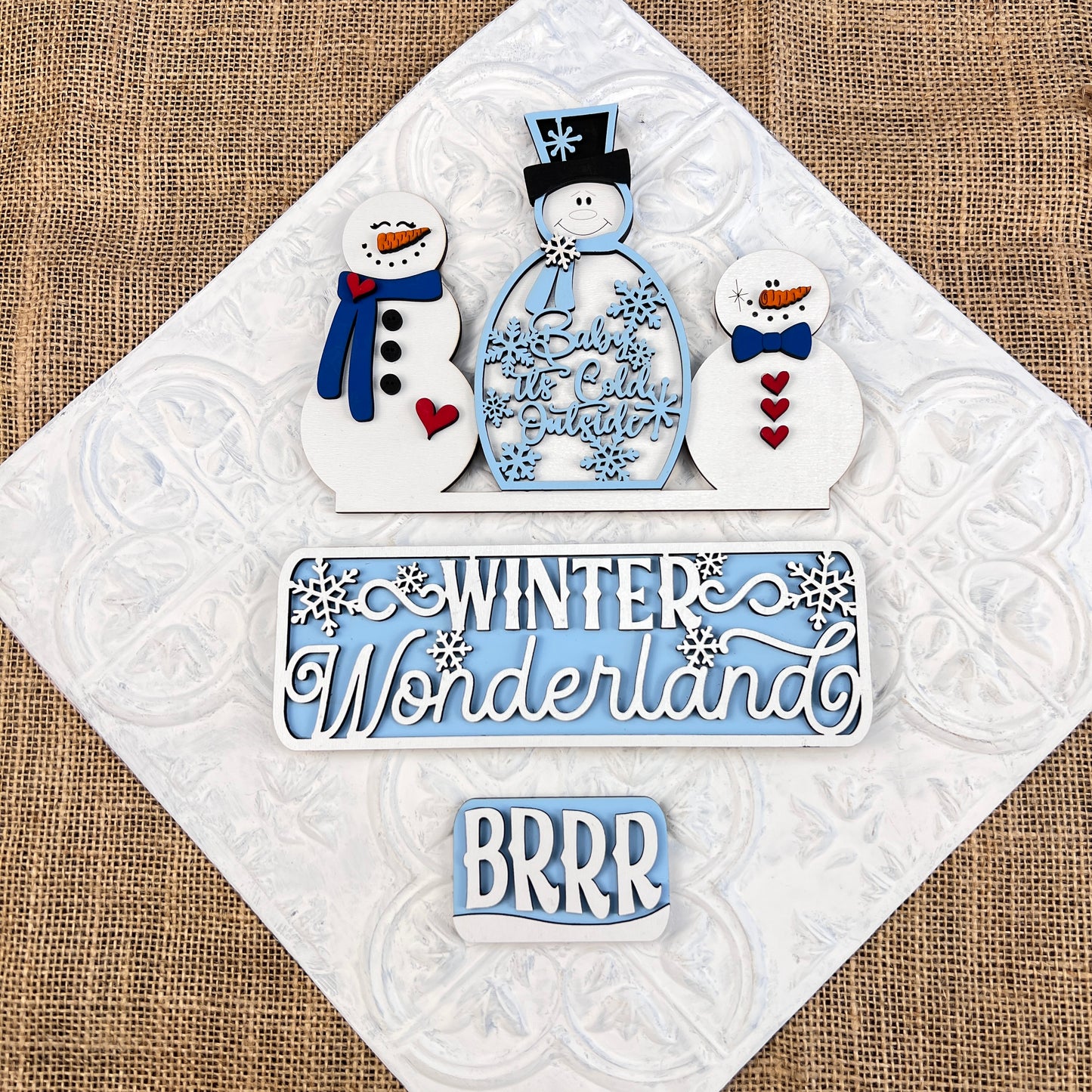 A set of hand-painted snowman cookies alongside a textured surface adorned with winter-themed signs from Janet's Craft Corner. These signs, crafted to resemble the "Winter Wonderland Interchangeable Insert - DIY Craft Kit," display festive blue and white messages like "Baby It's Cold Outside," "Winter Wonderland," and "BRRR.