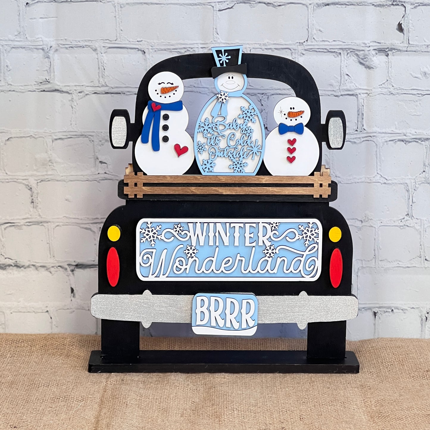 Janet's Craft Corner presents the "Winter Wonderland Interchangeable Insert - DIY Craft Kit - Set of 1," a decorative wooden sign featuring a truck with three snowmen dressed in winter attire. This home decor piece, perfect for creative enthusiasts, reads "Winter Wonderland" and "BRRR," and showcases snowflake accents against a brick wall and burlap surface.