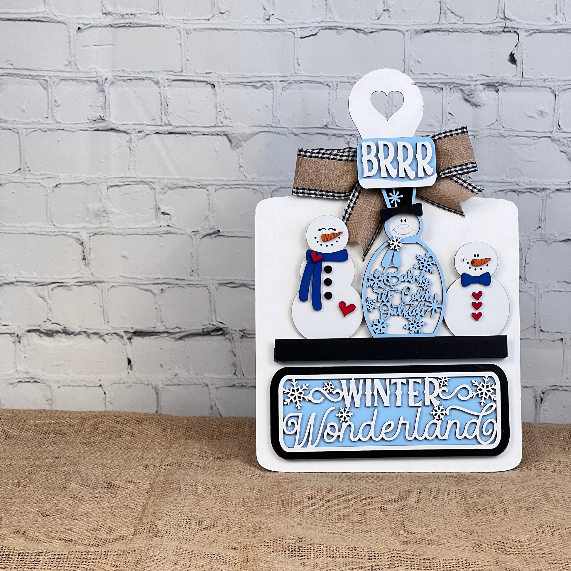 The Winter Wonderland Interchangeable Insert - DIY Craft Kit, presented by Janet's Craft Corner, allows you to craft a charming winter display featuring snowmen, hearts, and snowflakes. The scene is enhanced with phrases like "BRRR" and "Winter Wonderland" against a white brick wall backdrop on a wooden surface. This set is ideal for fans of interchangeable home decor.