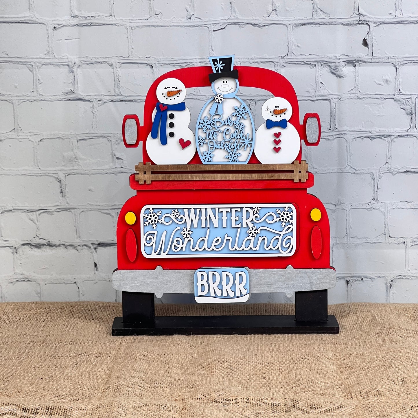 The Winter Wonderland Interchangeable Insert DIY Craft Kit by Janet's Craft Corner features a charming decorative piece depicting a red truck with two smiling snowmen. The tailgate is adorned with the words "Winter Wonderland" and "BRRR" in white and blue against a white brick wall, all placed on a burlap surface. It's perfect for interchangeable home decor!