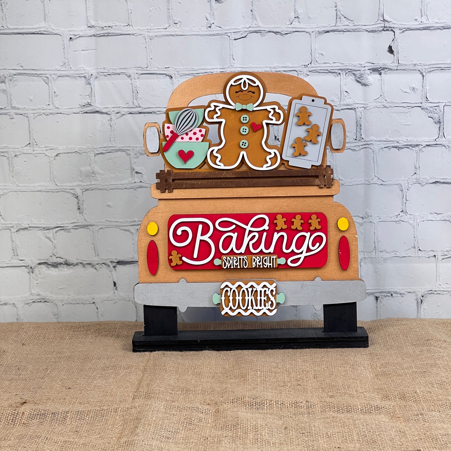 The Antique Truck Base with Insert DIY kit by Janet's Craft Corner features a wooden decoration with baking-themed items like a gingerbread man, cookie sheet, and whisk. It displays "Baking Spirits Bright" and "Cookies" against a white brick wall background, perfect for home décor enthusiasts!.