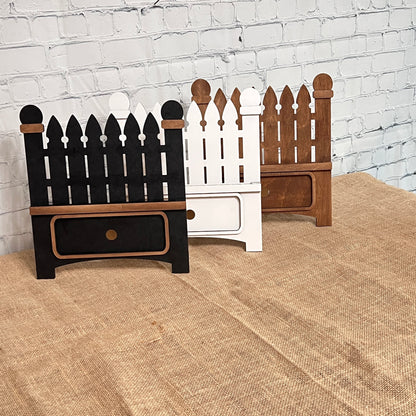 Three small picket fence-shaped decorations from Janet's Craft Corner rest on burlap against a white brick wall. These black, white, and brown DIY interchangeable pieces each have a drawer, perfect for personalized home décor.