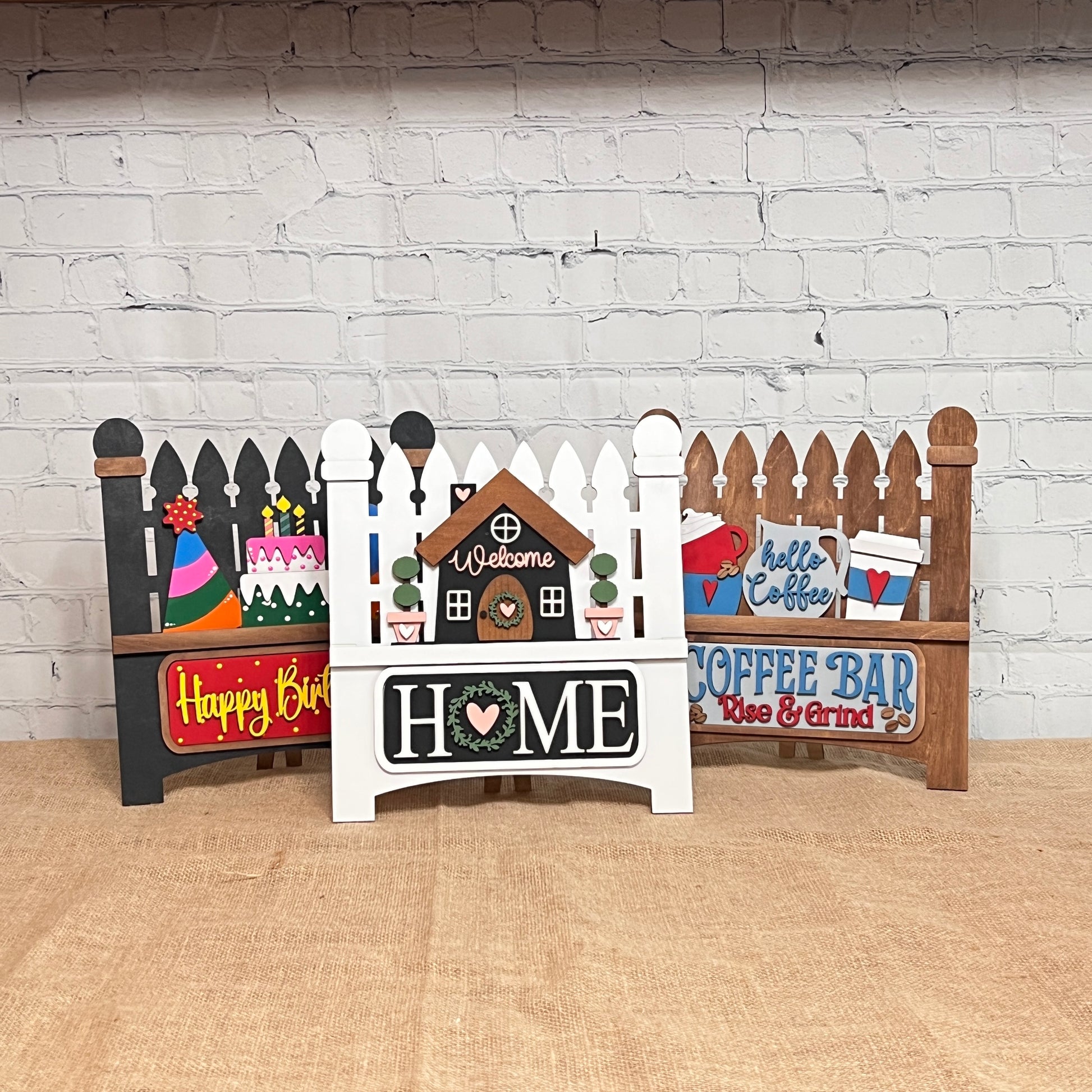 Janet's Craft Corner offers a Picket Fence with Insert DIY kit, perfect for home décor. Each set includes a fence base and one themed insert like "Welcome Home," "Happy Birthday," or "Coffee Bar." Hand-painted, they add charm to your space against any brick wall backdrop.