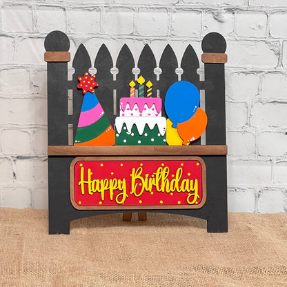 The Birthday Interchangeable Insert by Janet's Craft Corner features a wooden cake, candles, party hat, and balloons with "Happy Birthday" in yellow on a red background. Perfect for any birthday celebration, this DIY home decor craft kit includes a 3-piece set ideal for burlap surfaces against white brick walls.