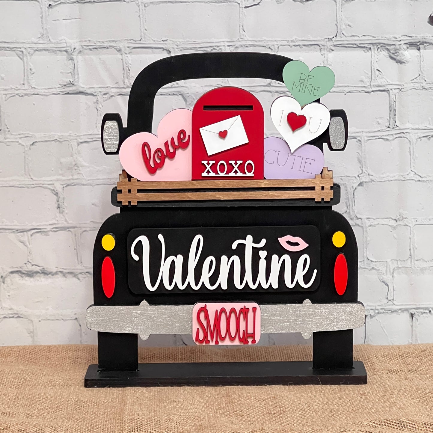 The Valentine Interchangeable Insert DIY home decor kit from Janet's Craft Corner includes a black truck cutout with "Valentine" on the tailgate. The truck bed features pastel hearts with sweet words, a red mailbox labeled "XOXO," and an interchangeable "smooch" sign against a white brick wall backdrop.