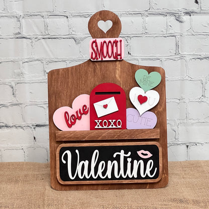 Janet's Craft Corner offers a Valentine Interchangeable Insert DIY home decor kit featuring a wooden board with a heart cutout, colorful hearts with messages like "love" and "cutie," an envelope, and a blackboard with "Valentine," perfect for creating personalized Valentine's Day displays.