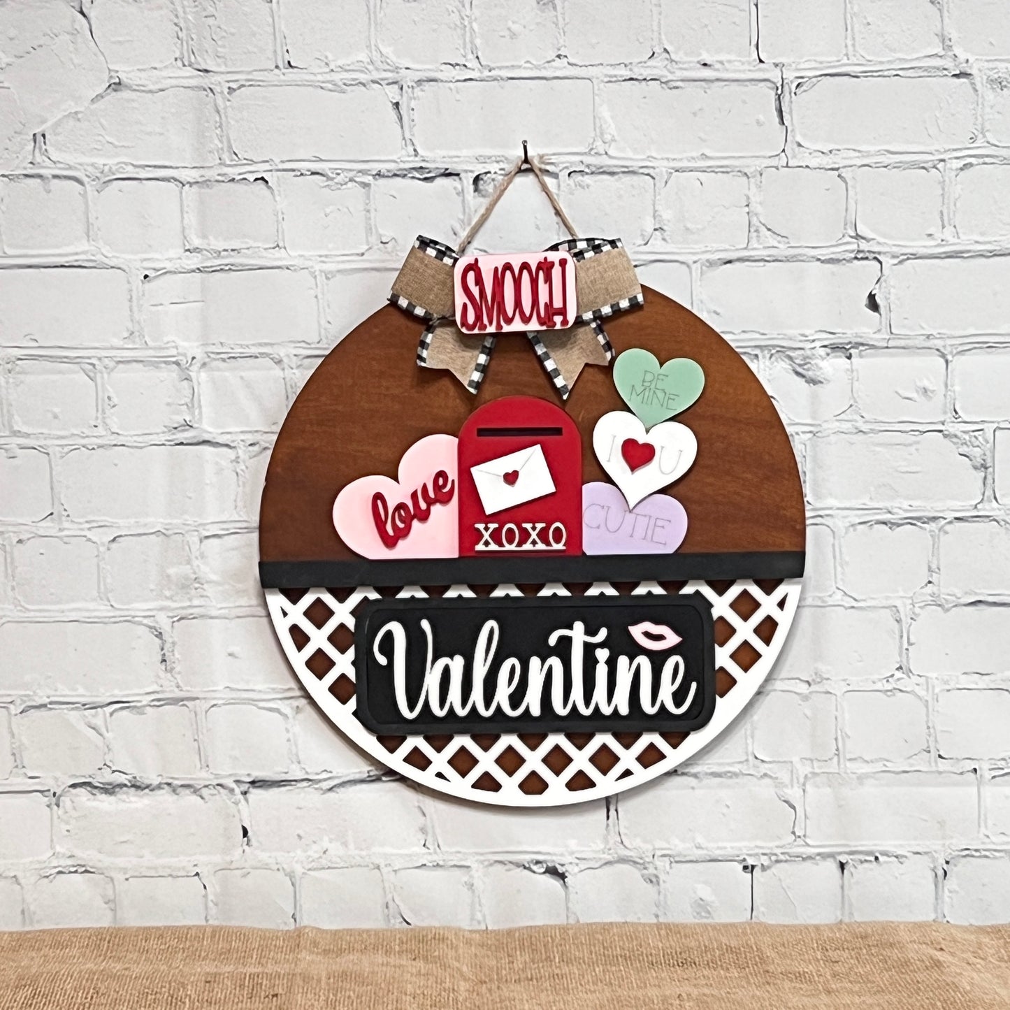 The Valentine Interchangeable Insert DIY home decor kit from Janet's Craft Corner includes a round hanging decoration with a bow, featuring a bold "Valentine" sign, hearts saying "Love," "XOXO," and "Be Mine," plus an image of a mailbox and a "Smooch" sign. Perfect for Valentine's Day crafting!.