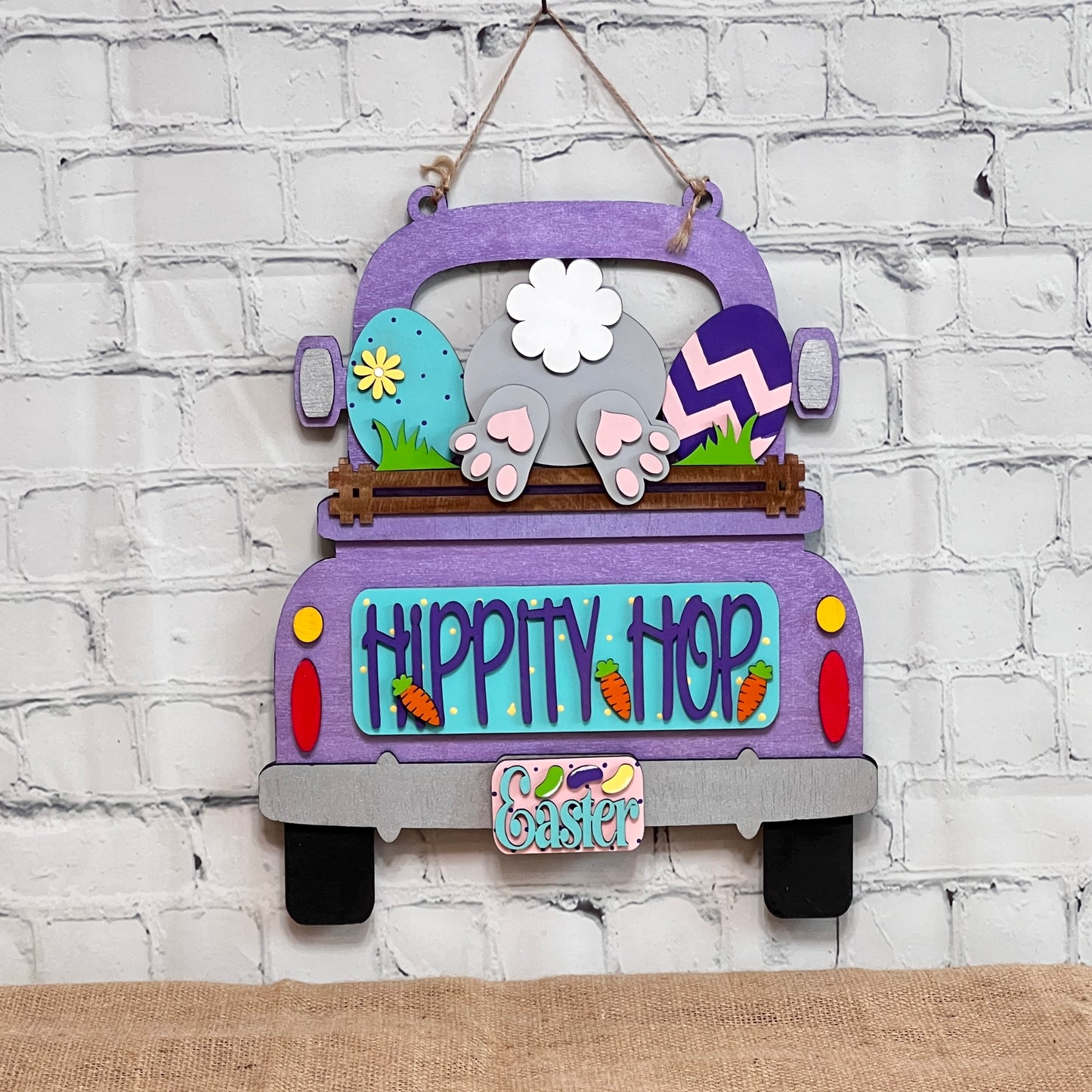 The "Hippity Hop Bunny Butt Interchangeable Insert" from Janet's Craft Corner features a hand-painted purple truck filled with Easter eggs and a bunny's hindquarters, set against a white brick background. It includes the text "Hippity Hop" with an "Easter" sign below, ideal for home decor.