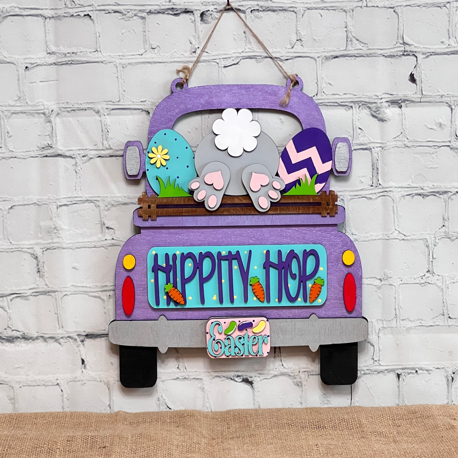 The "Hippity Hop Bunny Butt Interchangeable Insert" from Janet's Craft Corner features a hand-painted purple truck filled with Easter eggs and a bunny's hindquarters, set against a white brick background. It includes the text "Hippity Hop" with an "Easter" sign below, ideal for home decor.