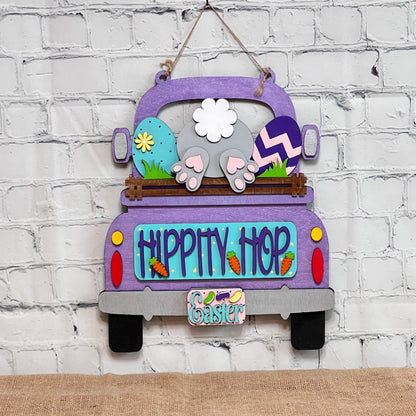 The "Hippity Hop Bunny Butt Interchangeable Insert" from Janet's Craft Corner features a hand-painted purple truck filled with Easter eggs and a bunny's hindquarters, set against a white brick background. It includes the text "Hippity Hop" with an "Easter" sign below, ideal for home decor.