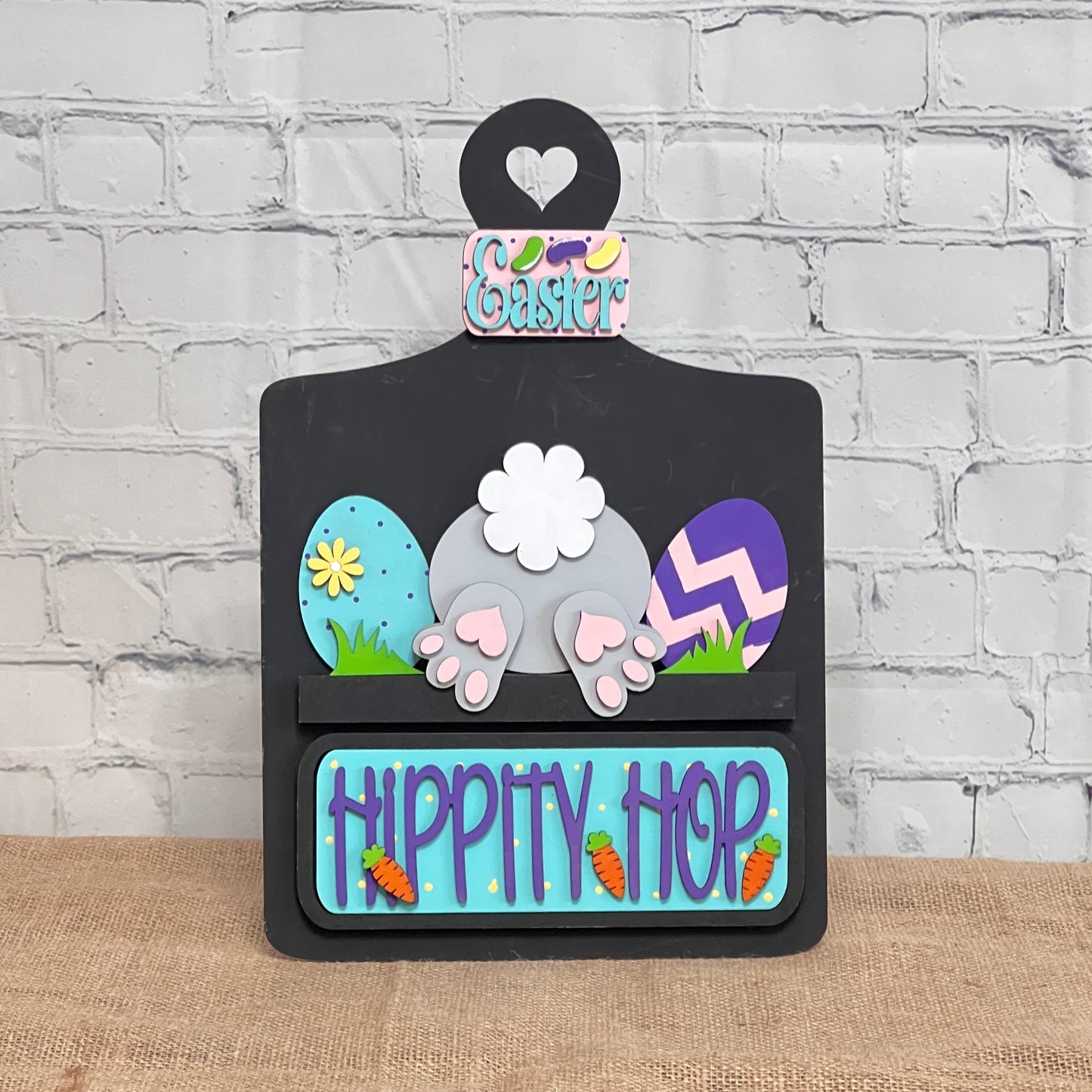 The "Hippity Hop Bunny Butt Interchangeable Insert" by Janet's Craft Corner features a decorative bunny rear with Easter eggs. The "Hippity Hop" sign is perfect home decor against a white brick wall for a festive touch.