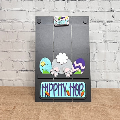 The Hippity Hop Bunny Butt Interchangeable Insert by Janet's Craft Corner features colorful eggs, bunny ears, a fluffy tail, "Hippity Hop" text, and carrot illustrations on a decorative Easter sign. This charming hand-painted piece enhances any home decor collection.