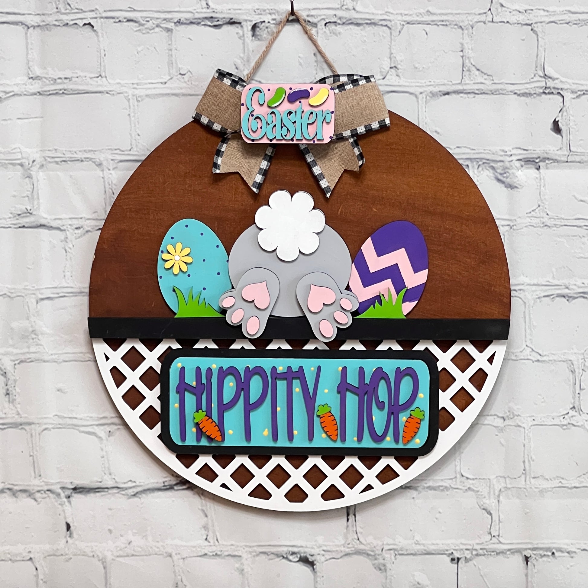 The DIY craft kit "Hippity Hop Bunny Butt Interchangeable Insert" by Janet's Craft Corner features a hand-painted wooden bunny's back, surrounded by colorful eggs and small carrots. The sign reads "Hippity Hop," and it hangs on a brick wall with an "Easter" bow at the top.
.