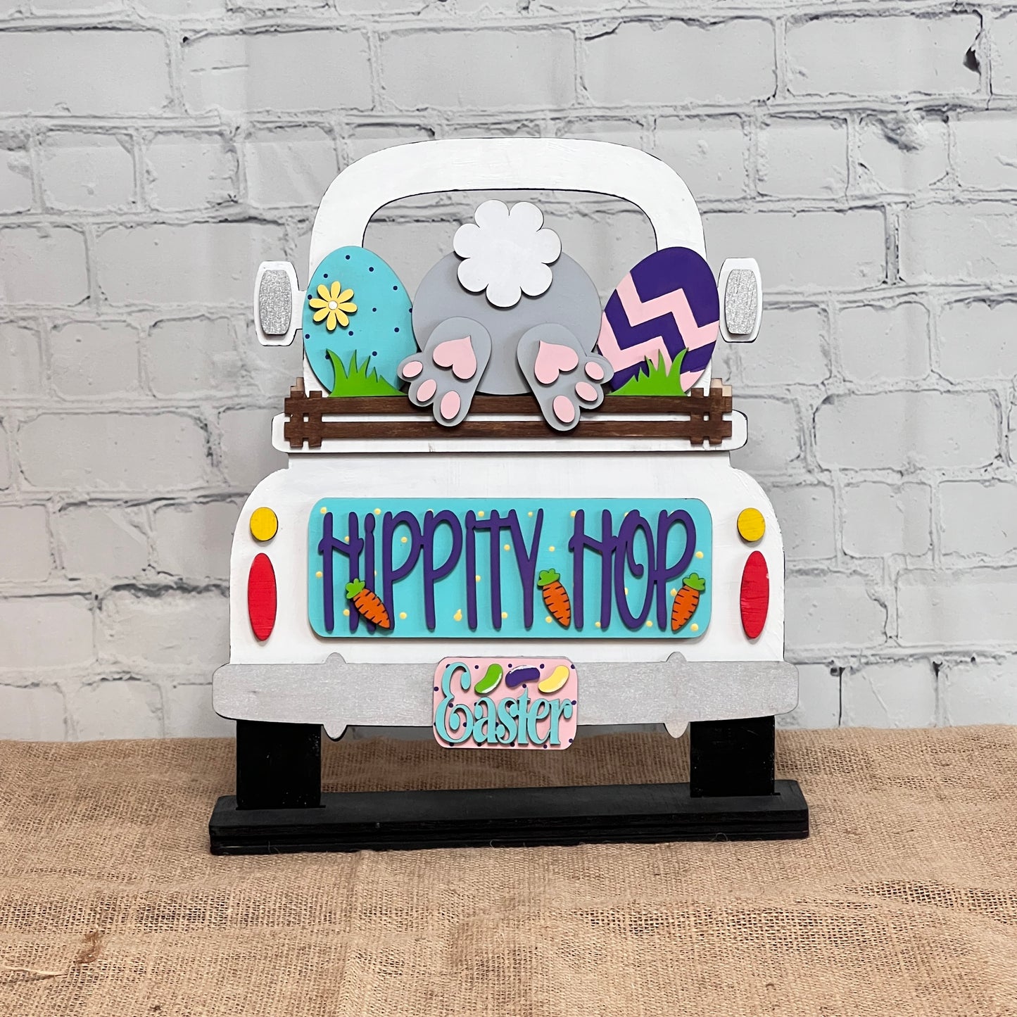 The "Hippity Hop Bunny Butt Interchangeable Insert" by Janet's Craft Corner is a decorative sign featuring a hand-painted bunny tail and feet in a truck bed, surrounded by Easter eggs, flowers, and carrot decorations against a brick wall backdrop for versatile home decor.