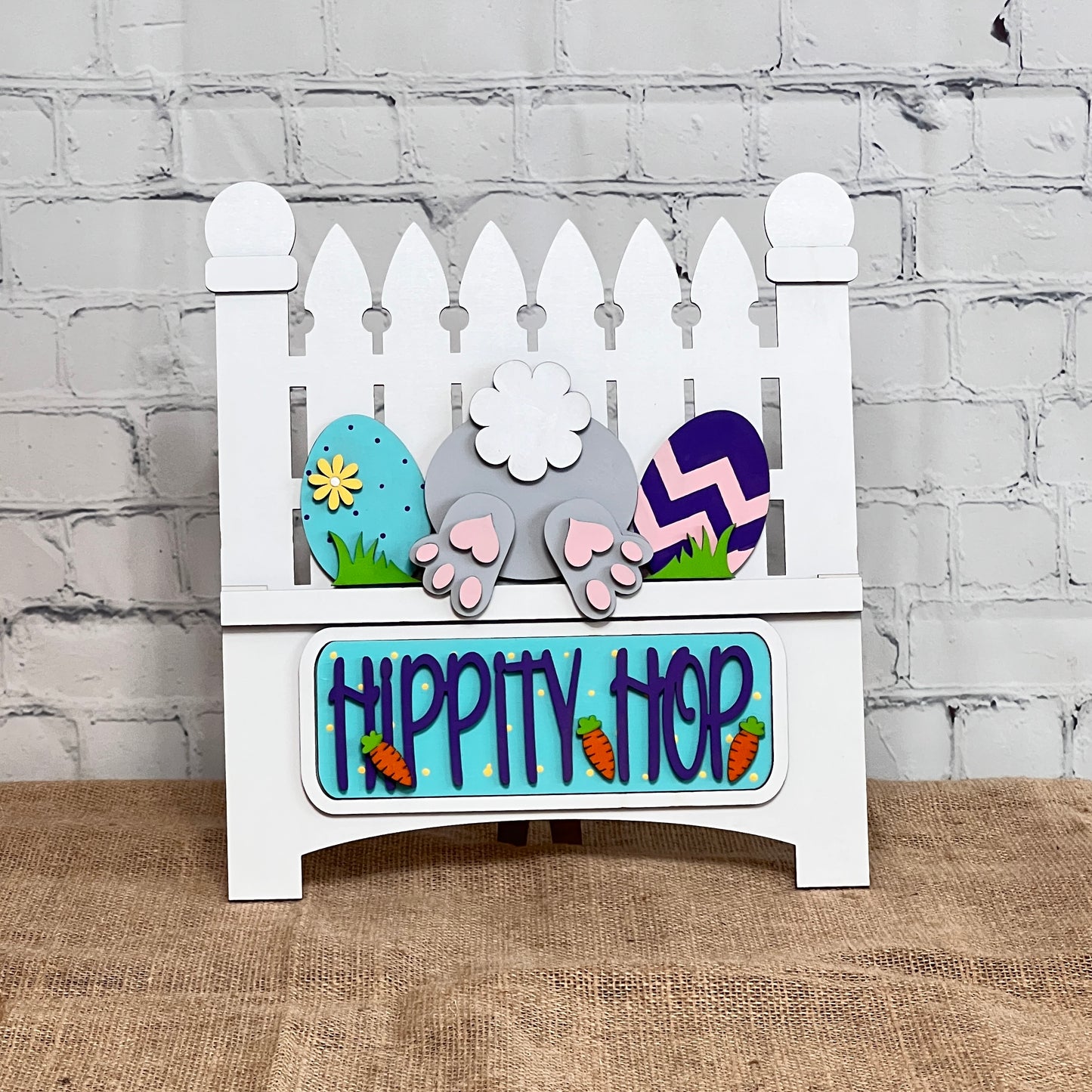 The Hippity Hop Bunny Butt Interchangeable Insert from Janet's Craft Corner is a charming DIY craft kit featuring a decorative white picket fence sign with a bunny tail, colorful eggs, paw prints, and "Hippity Hop" in blue with carrot accents on burlap against a white brick wall.