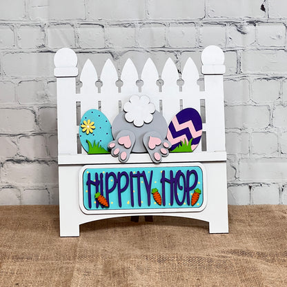 The Hippity Hop Bunny Butt Interchangeable Insert from Janet's Craft Corner is a charming DIY craft kit featuring a decorative white picket fence sign with a bunny tail, colorful eggs, paw prints, and "Hippity Hop" in blue with carrot accents on burlap against a white brick wall.