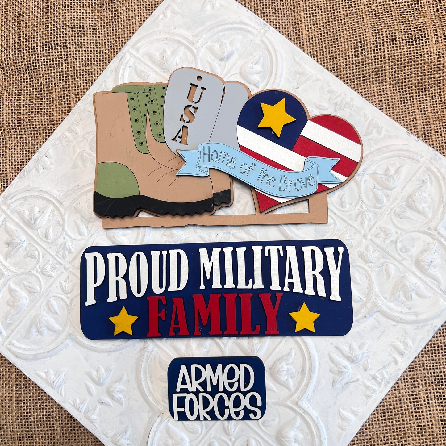 The Military Interchangeable Insert by Janet's Craft Corner features a boot, "USA" dog tags, a heart with stars and stripes, the text "Home of the Brave," and plaques reading "Proud Military Family" and "Armed Forces." Ideal for interchangeable home decor or as a DIY hand-painted craft kit.