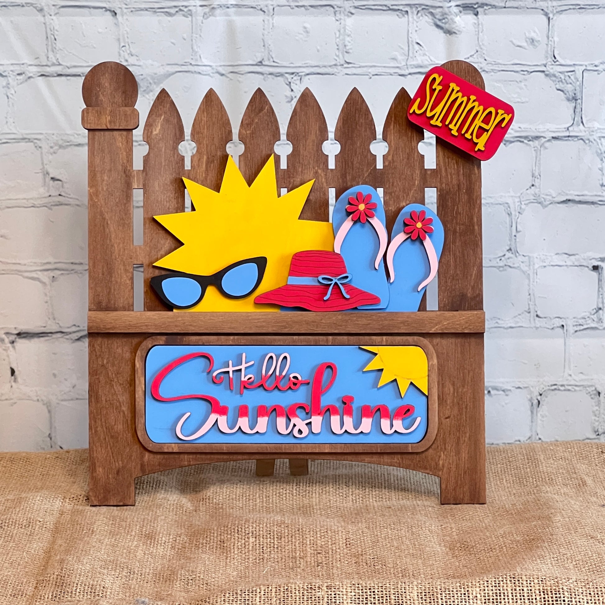 The Hello Sunshine Interchangeable Insert by Janet's Craft Corner is a DIY seasonal decor kit. It includes a picket fence design featuring a sun with sunglasses and a red hat, flip-flops, "Hello Sunshine" text, and a mini “Summer Picnic” sign against a white brick wall backdrop on burlap.