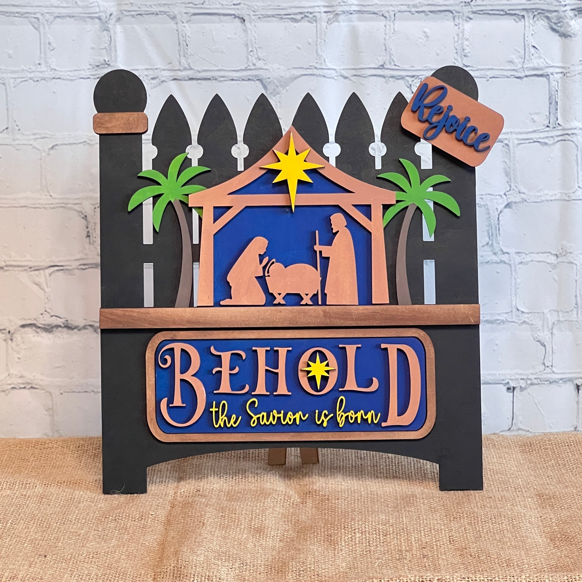 The Nativity Interchangeable Insert from Janet's Craft Corner is a DIY seasonal home decor craft kit featuring silhouettes of Mary, Joseph, and baby Jesus under a star, with "Behold the Savior is born" text in blue and orange. Palm trees flank the scene against a festive brick wall background.