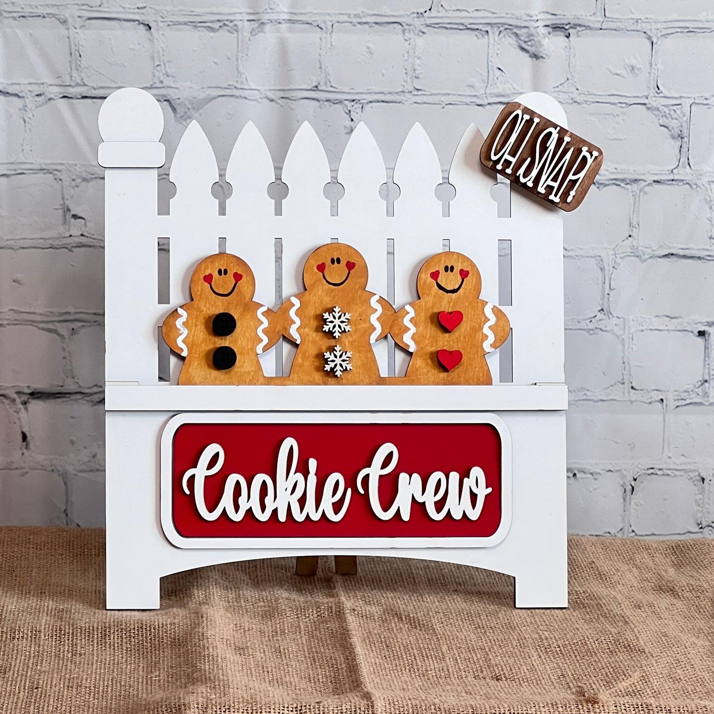 The Cookie Crew Interchangeable Insert by Janet's Craft Corner is a DIY home decor craft kit featuring three uniquely patterned gingerbread men on a white picket fence. The board reads "Oh Snap!" and includes a red "Cookie Crew" sign against a white brick wall, perfect for seasonal inserts.