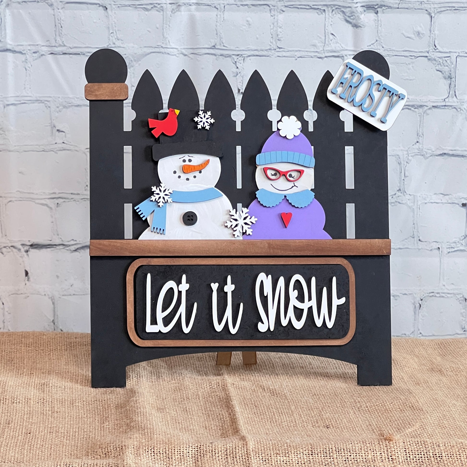 The "Let It Snow" Interchangeable Insert by Janet's Craft Corner is a DIY craft kit that creates a three-piece decorative wooden sign featuring a picket fence with a snowman, a purple-capped figure, and a cardinal against a brick wall. The insert reads "Frosty.