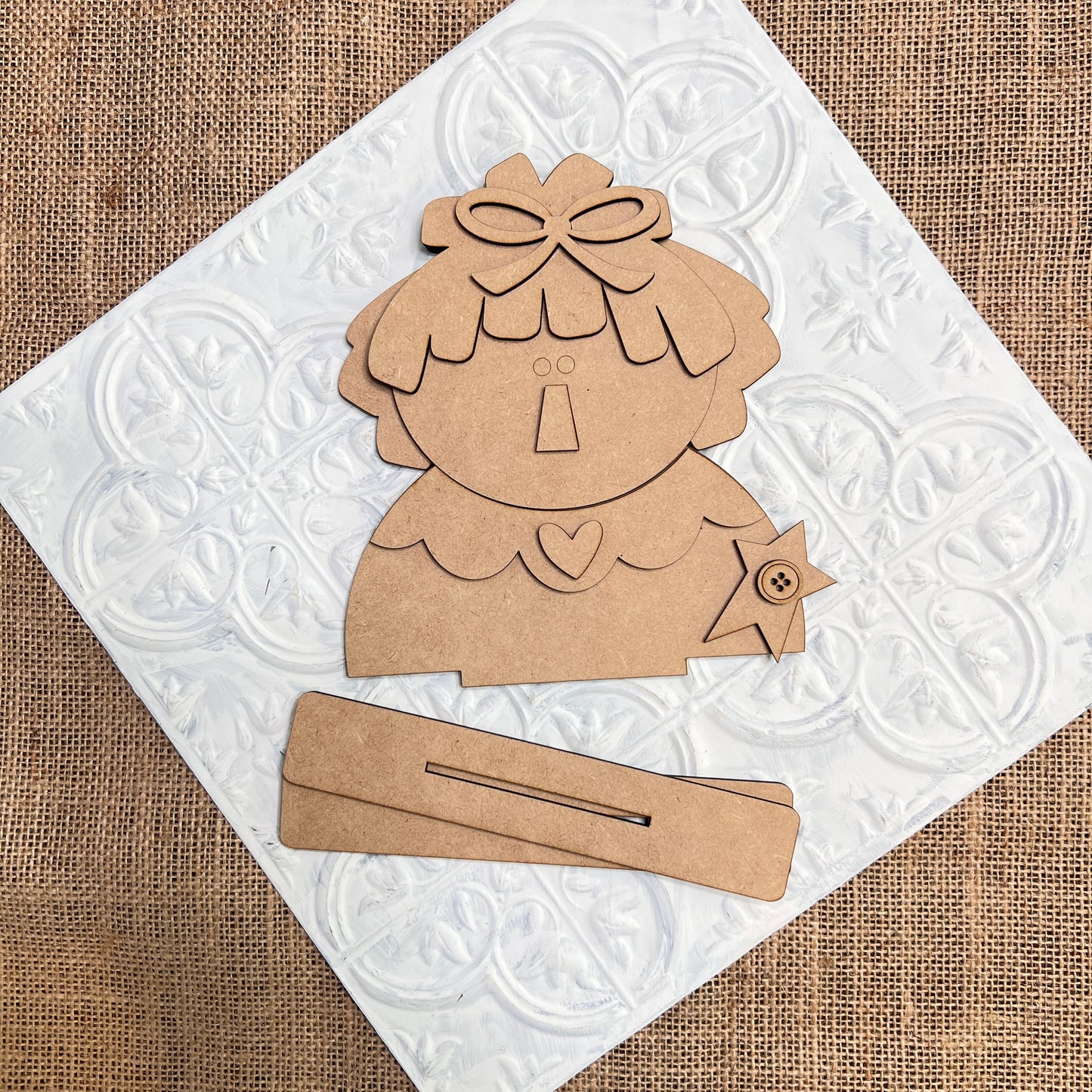 Janet's Craft Corner’s Raggedy Ann & Andy Shelf Sitters DIY kit features whimsical cardboard pieces with bow, heart, and star accents, displayed on white tile over burlap for vintage charm in your seasonal home decor creations.