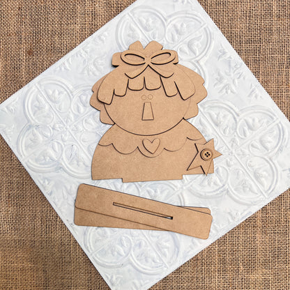 Janet's Craft Corner’s Raggedy Ann & Andy Shelf Sitters DIY kit features whimsical cardboard pieces with bow, heart, and star accents, displayed on white tile over burlap for vintage charm in your seasonal home decor creations.