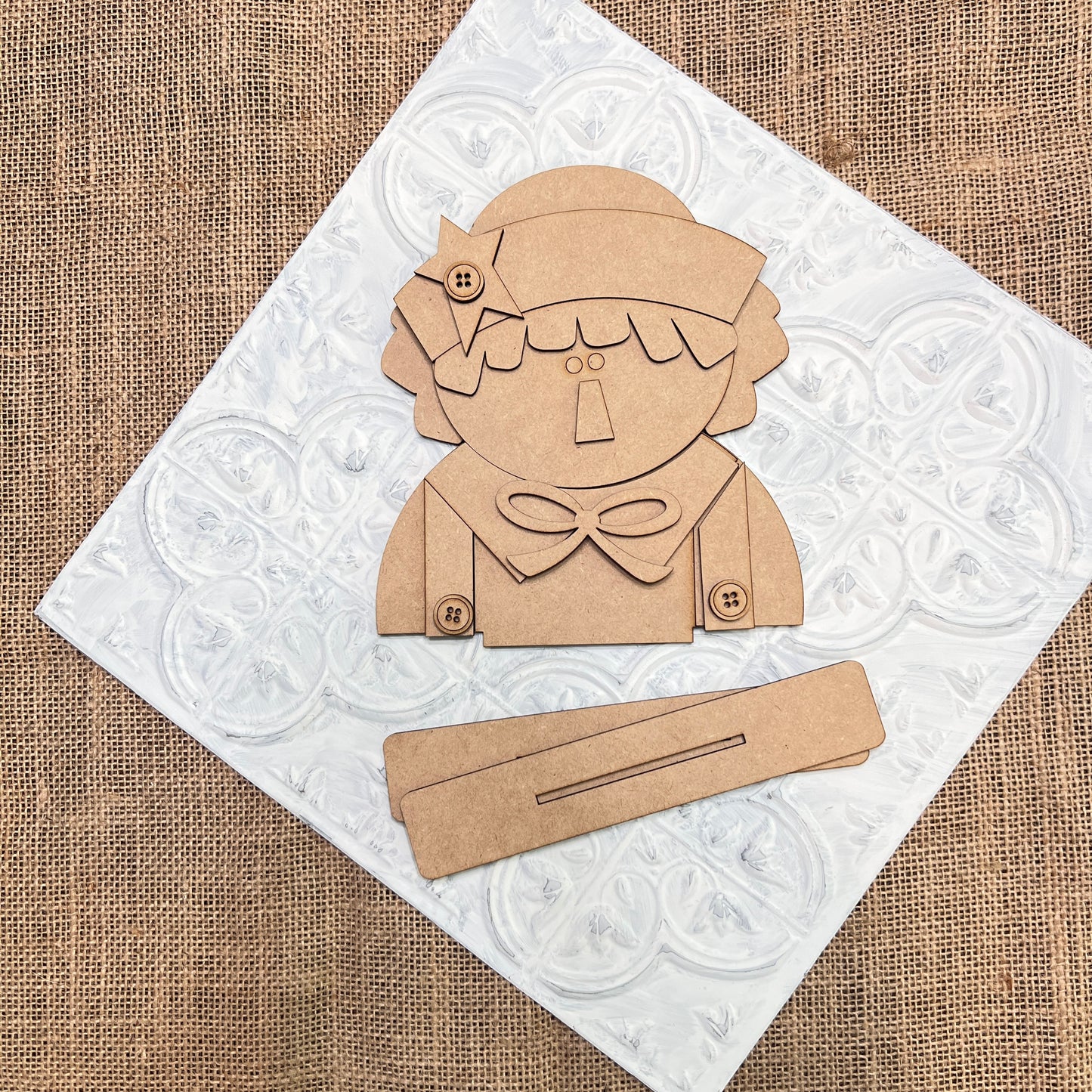 A flat lay of Janet's Craft Corner Raggedy Ann & Andy Shelf Sitters DIY seasonal home decor craft kit set, featuring a brown cut-out figure with a bow and buttons on a white tile over burlap, plus two rectangular pieces in front.