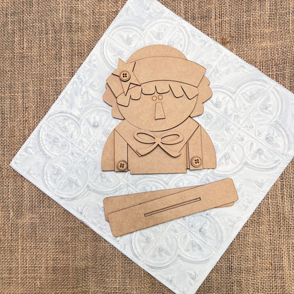 A flat lay of Janet's Craft Corner Raggedy Ann & Andy Shelf Sitters DIY seasonal home decor craft kit set, featuring a brown cut-out figure with a bow and buttons on a white tile over burlap, plus two rectangular pieces in front.