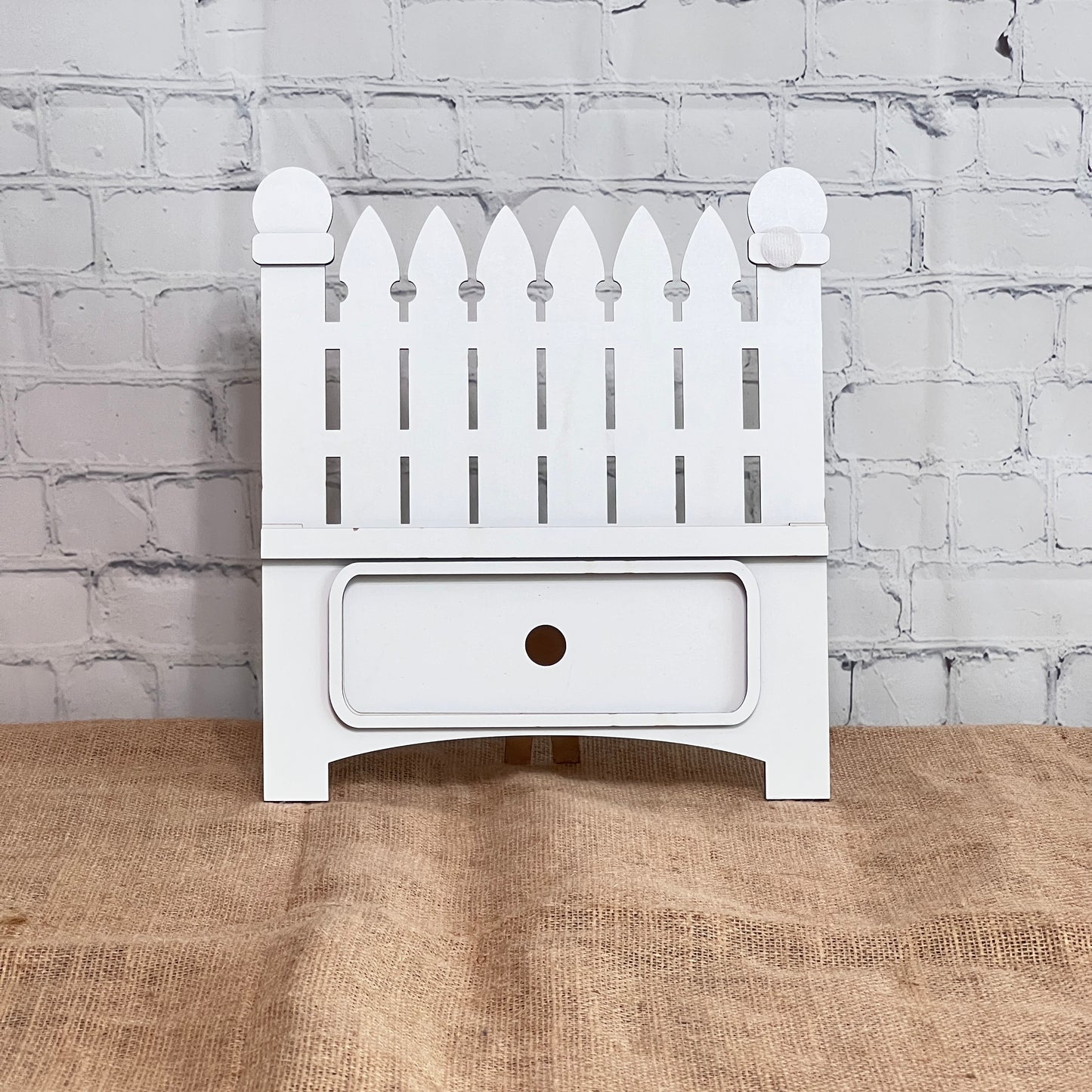 The Garden Fence Interchangeable Base - Painted from Janet's Craft Corner is a small white drawer unit featuring a picket fence craft kit on top, set against a white brick wall and brown burlap—ideal for customizable DIY home decor.