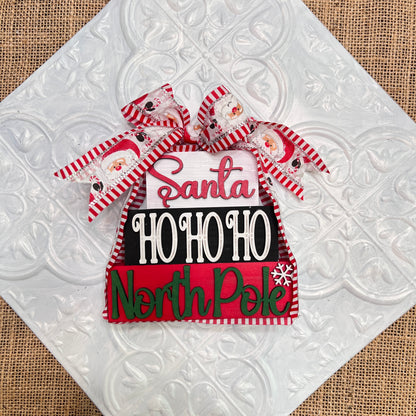 Janet’s Craft Corner’s Word Stacks - Painted is a festive Christmas sign with ribbons, featuring “Santa,” “HO HO HO,” and “North Pole” in red, white, and green on a textured white tile background.