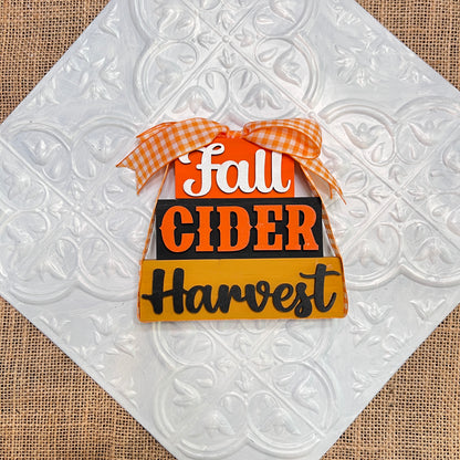 Janet's Craft Corner Word Stacks - Painted: A decorative candy corn-shaped sign with an orange gingham bow and "Fall Cider Harvest" in white, orange, and black letters, shown on patterned tile with burlap underneath.