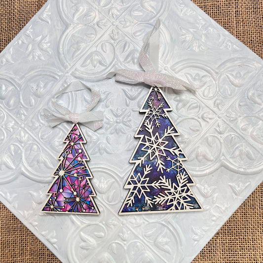 Two hand-cut Tree Ornaments from Janet’s Craft Corner rest on a textured white tile. One ornament is pink with a geometric design; the other is blue and purple, featuring delicate snowflake patterns. Both include ribbons for hanging.