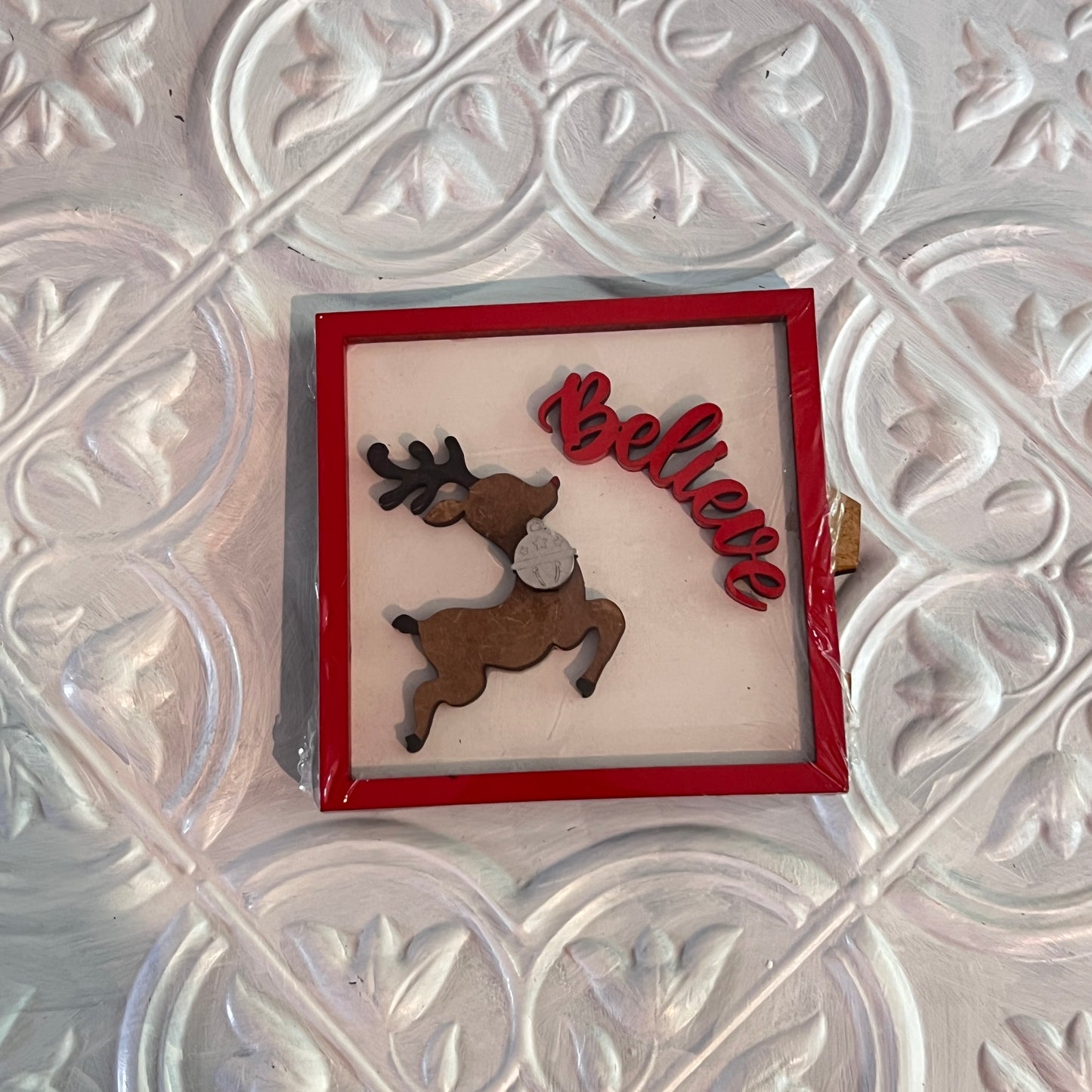 Janet's Craft Corner Christmas Mini Shelf Sitters: This square sign has a red border, displays a leaping reindeer, and "Believe" in red cursive atop a white textured decorative background.
