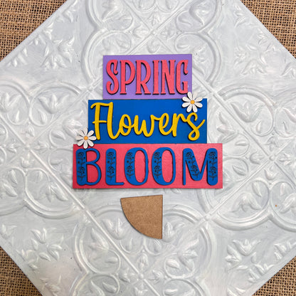 The Word Stacks - Painted by Janet's Craft Corner features "SPRING Flowers BLOOM" in colorful, layered text with two white daisies, displayed on a white patterned tile background.