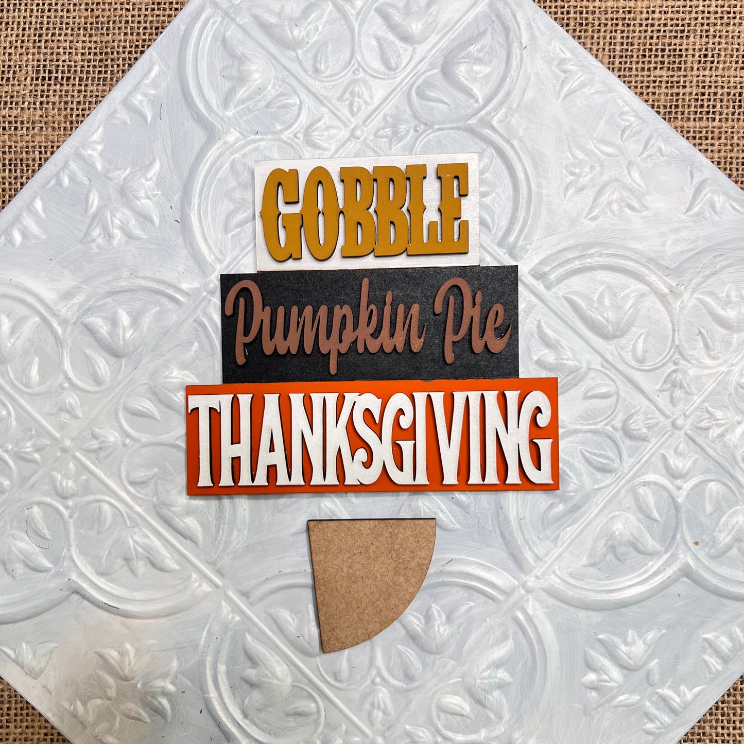 The Word Stacks - Painted sign by Janet's Craft Corner features "Gobble," "Pumpkin Pie," and "Thanksgiving" stacked above a brown pie-shaped cutout on a textured white background.
