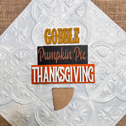 The Word Stacks - Painted sign by Janet's Craft Corner features "Gobble," "Pumpkin Pie," and "Thanksgiving" stacked above a brown pie-shaped cutout on a textured white background.