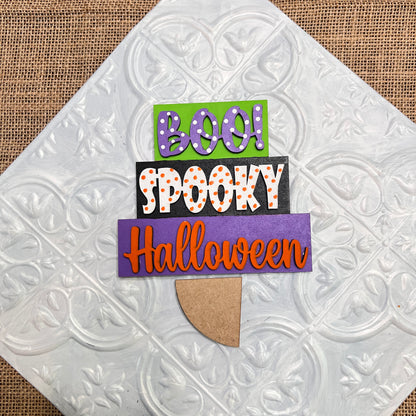 The Word Stacks - Painted sign by Janet's Craft Corner features "BOO!" in green, "SPOOKY" in white with orange dots, and "Halloween" in orange on a purple background, displayed on a white textured tile atop burlap.
