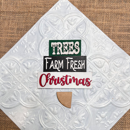 The Word Stacks - Painted sign from Janet's Craft Corner features "TREES" in green, "Farm Fresh" in white on black, and "Christmas" in red on a textured white background with a tan wood accent at the bottom.