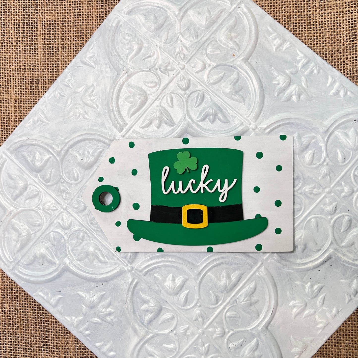 A 6 inch square from Janet's Craft Corner featuring a green leprechaun hat, "lucky" in white script, and green polka dots on a white textured tile over burlap.