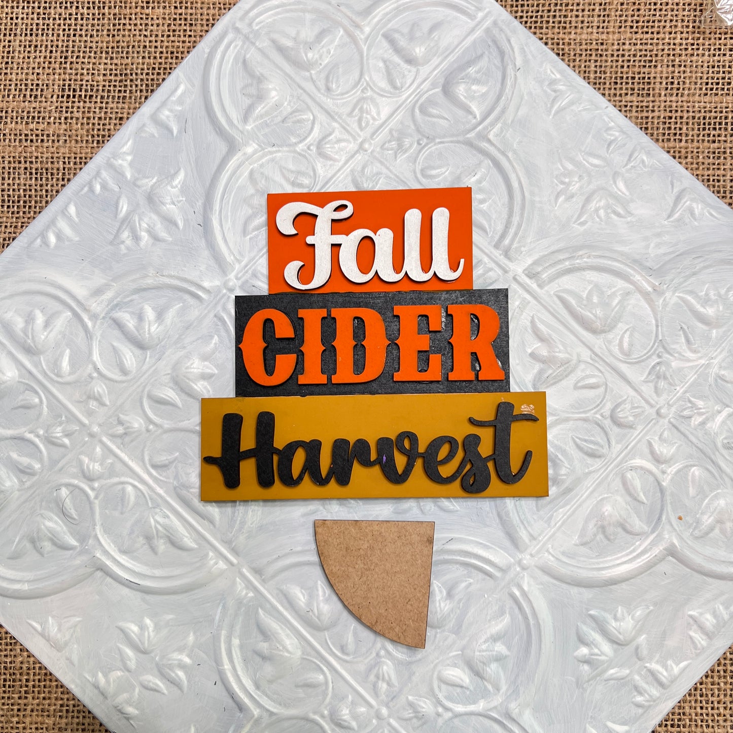 The Word Stacks - Painted by Janet's Craft Corner features "Fall" in white on orange, "Cider" in black on gray, and "Harvest" in black on mustard yellow, all arranged on a textured white tile with a small brown half-circle base.