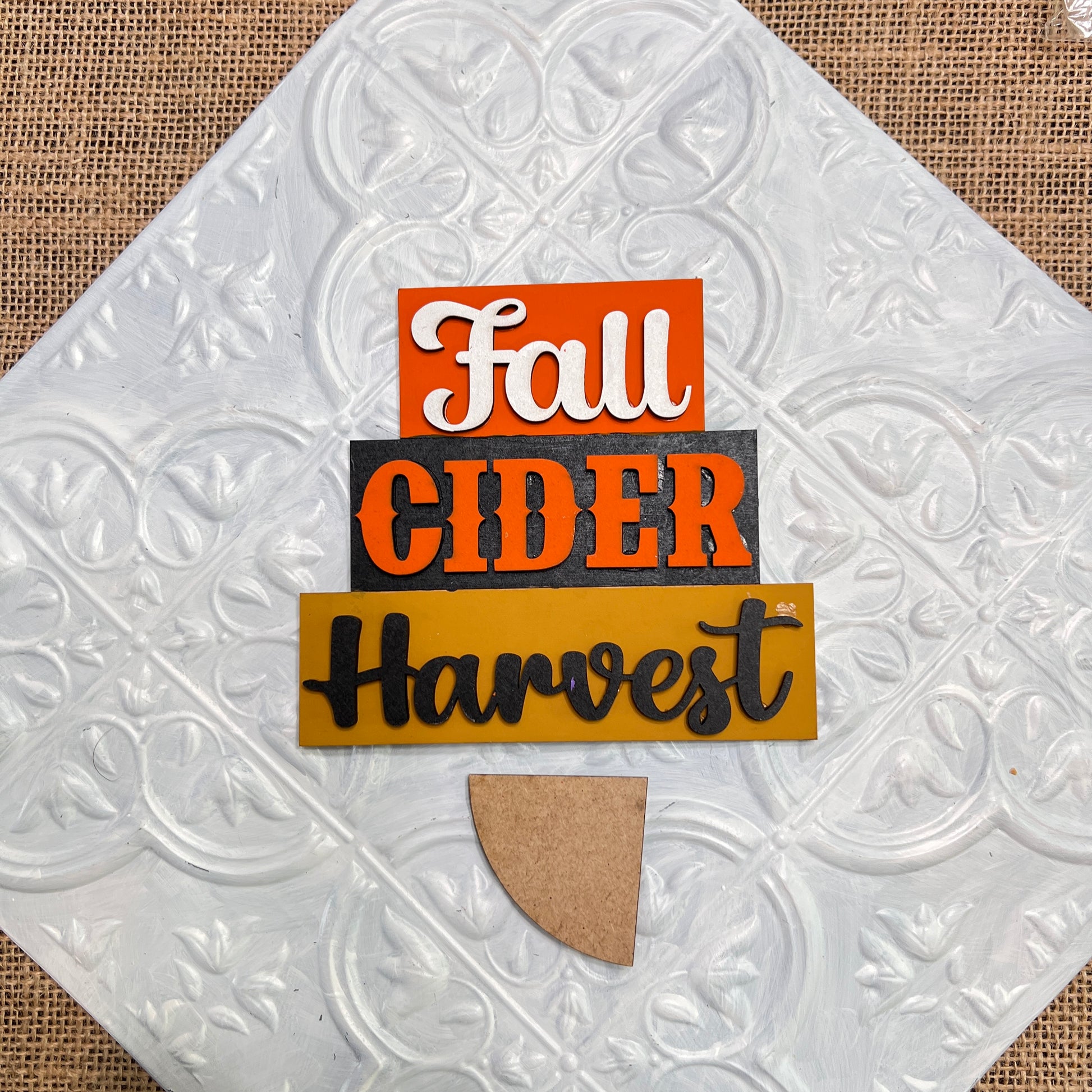 The Word Stacks - Painted by Janet's Craft Corner features "Fall" in white on orange, "Cider" in black on gray, and "Harvest" in black on mustard yellow, all arranged on a textured white tile with a small brown half-circle base.