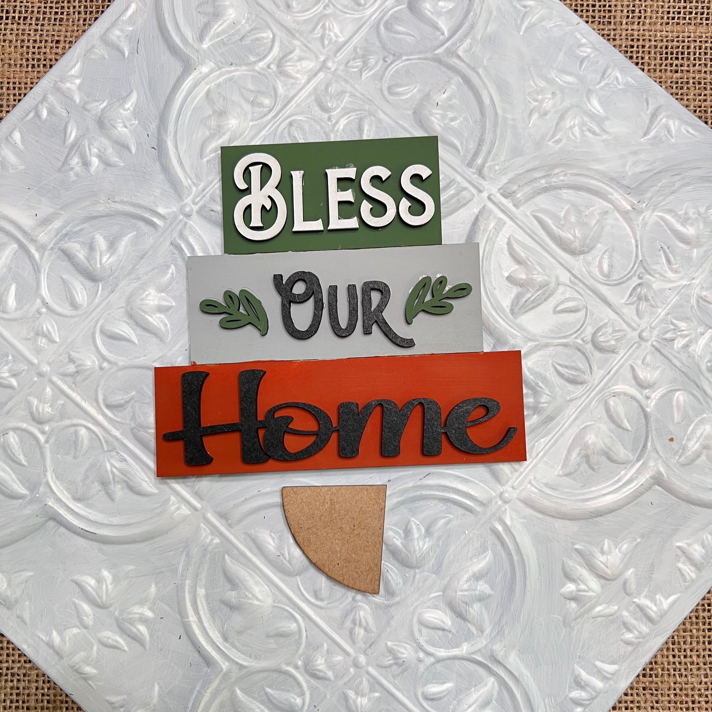 Janet's Craft Corner Word Stacks - Painted: A tree-shaped decorative sign with "Bless Our Home" on green, gray, and red planks and a small brown base, displayed on a textured white tile background.